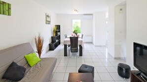 Uma sala de estar moderna com sofá, apoio para os pés, televisão de tela plana, área de jantar com cadeiras e mesa, e uma estante.