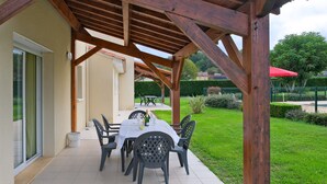 Alpendre coberto com mesa e cadeiras, pérgola de madeira e vista para um jardim bem cuidado.