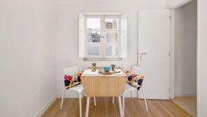 Un petit coin repas avec une table en bois et deux chaises blanches, une fenêtre au-dessus de la table et une porte sur la droite.