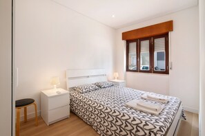 A bedroom with a bed, two bedside tables, a nightstand, a window with blinds, and a wooden floor.