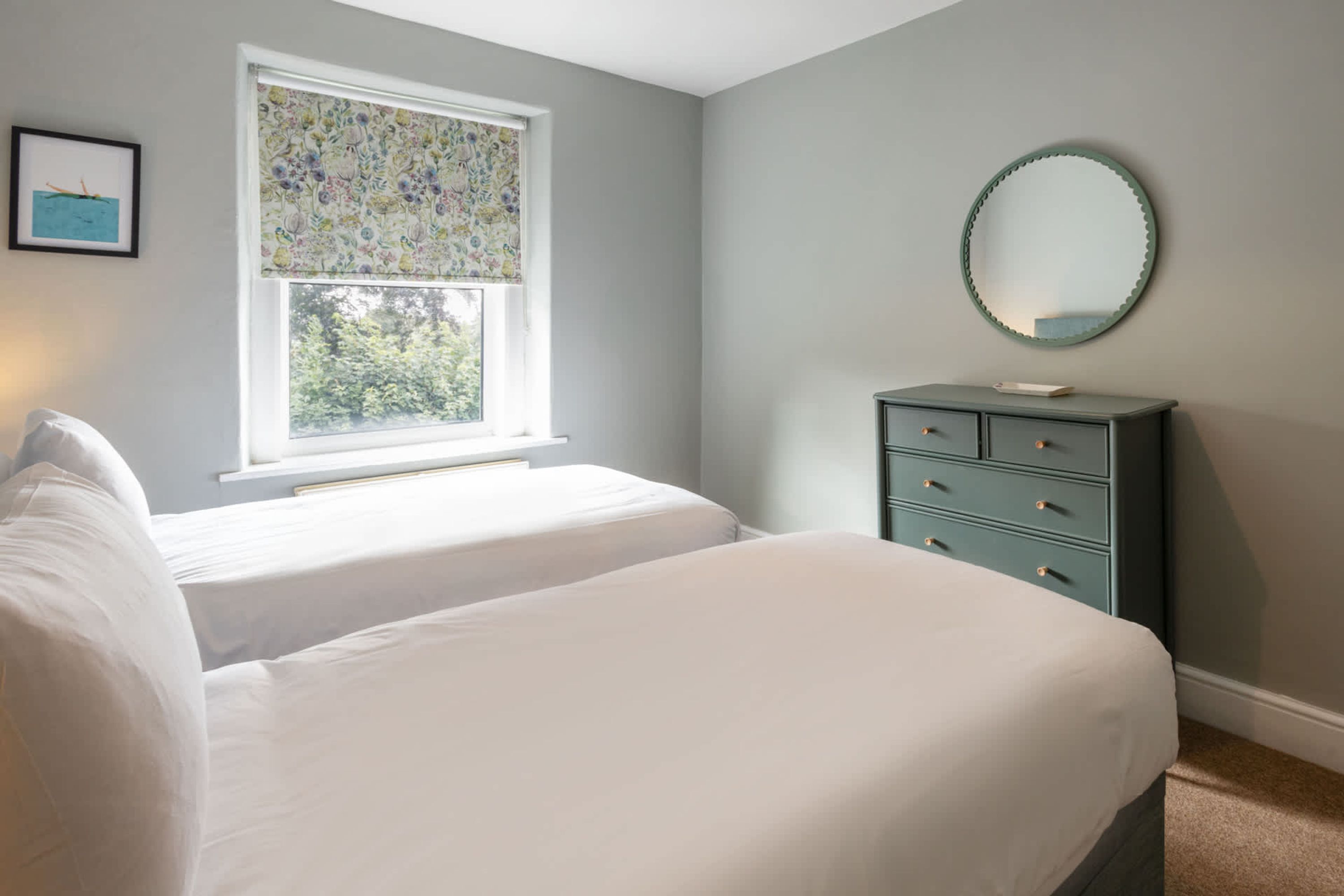 Une chambre avec deux lits, une commode, une fenêtre ornée de rideaux à motifs floraux et un miroir au mur.