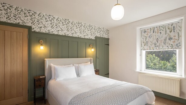 A bedroom with a bed, a nightstand, a window with a view, and a wall with a leafy pattern. - Host Stay Castle Hill Cottage (Keswick)