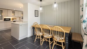 A modern kitchen with white cabinets, a central island, and a dining area with wooden chairs and a table. - Host Stay Castle Hill Cottage (Keswick)