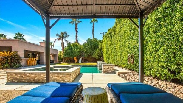 A covered outdoor area with lounge chairs, a pool, and a view of a modern house and palm trees. - Beautiful and Private 4 Bedroom Private Pool Home (Bermuda Dunes)