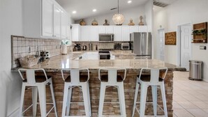 A modern kitchen with white cabinets, a large island with bar stools, stainless steel appliances, and decorative items on the shelves. - Beautiful and Private 4 Bedroom Private Pool Home (Bermuda Dunes)