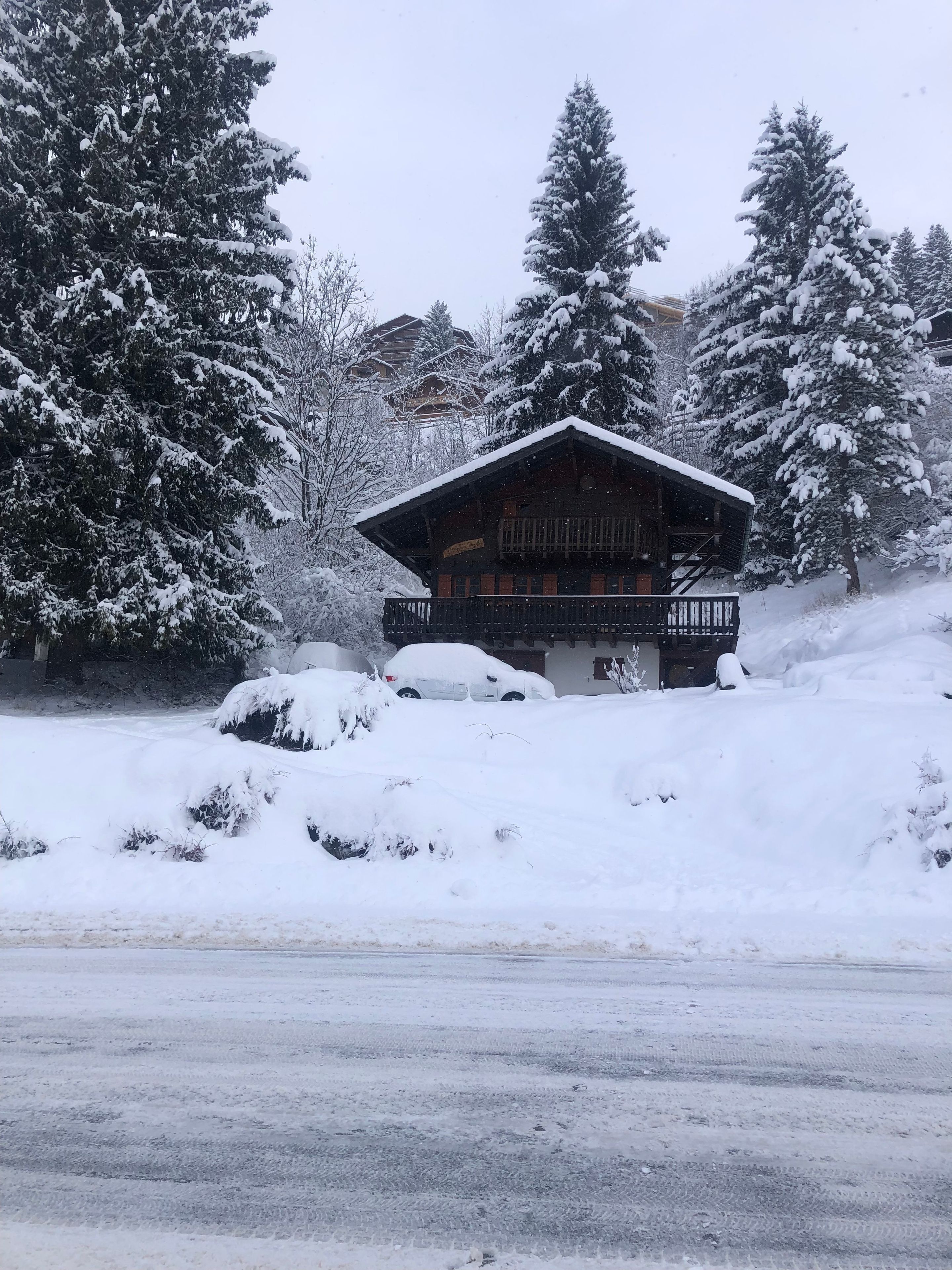 Un chalet enneigé, avec une façade en bois, un balcon et une voiture garée devant.