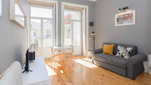 A living room with a grey sofa, a white table, and two chairs. There is a window with a view of buildings outside. - Liiiving in Porto - Silver Light Apartment (Porto)