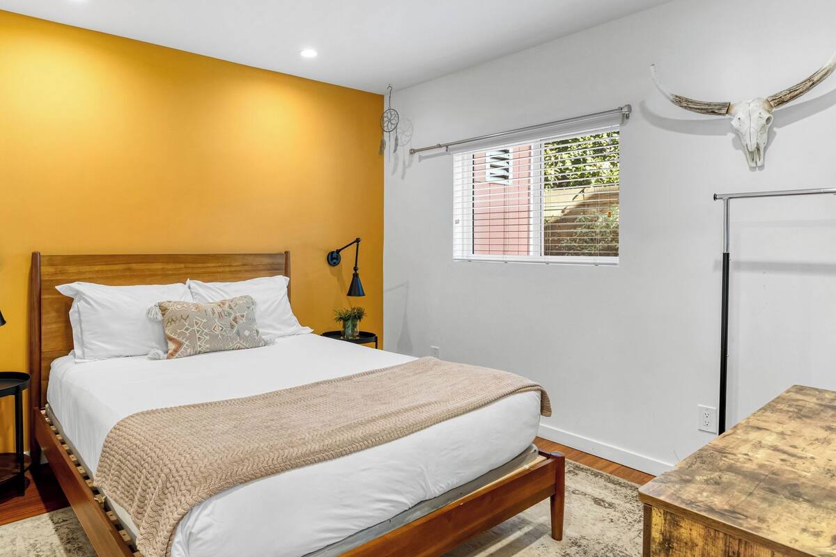 Un dormitorio con cama de madera, ropa de cama blanca, una mesita de noche pequeña, una lámpara de pared, una ventana con persianas y un cráneo decorativo en la pared.