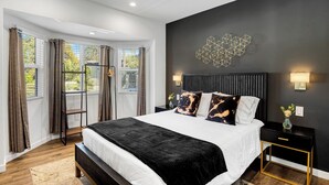 Quarto moderno com uma cama grande, parede escura com luminária geométrica e vista para o verde pelas janelas.