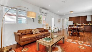 Un salon moderne avec un canapé en cuir marron, une table basse sur laquelle repose un livre, un lampadaire et un coin repas avec des chaises et une table en bois.