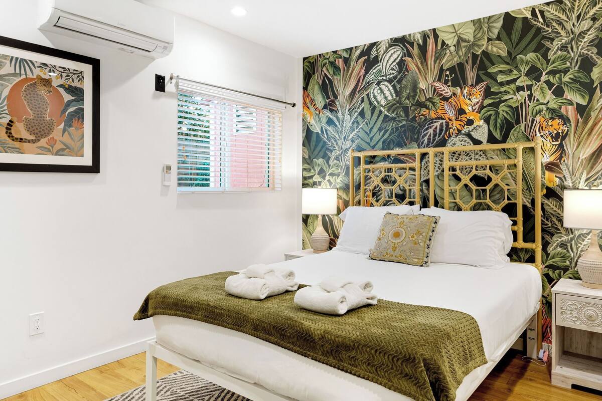 Un dormitorio con cama, dos mesitas de noche, una ventana con persianas, un aire acondicionado de pared y un cuadro enmarcado.