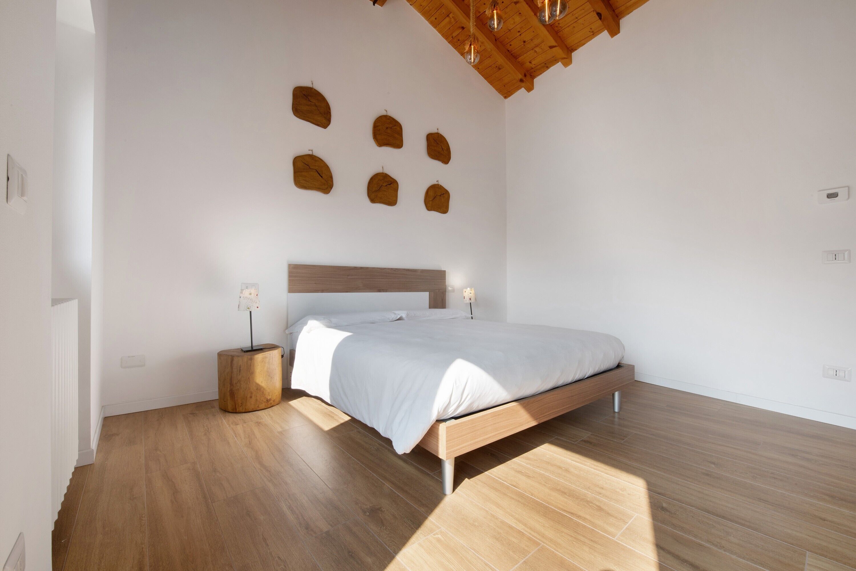 Ein Schlafzimmer mit einem hölzernen Bett, einem runden Nachttisch aus Holz, einem weißen Kopfteil und einer Holzbalkendecke.