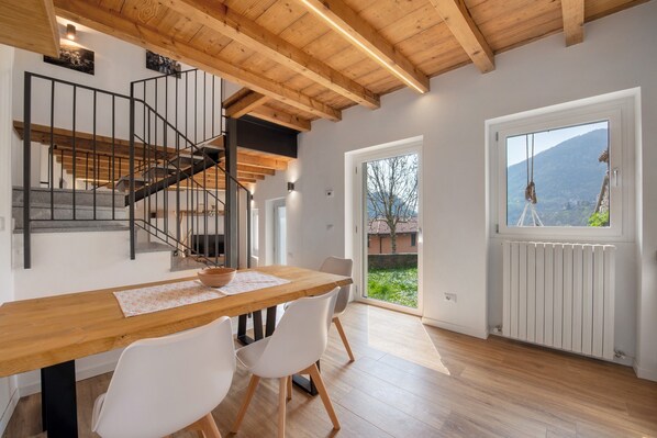 Un coin repas avec une table en bois, des chaises blanches, un escalier et une vue à travers une porte vitrée.