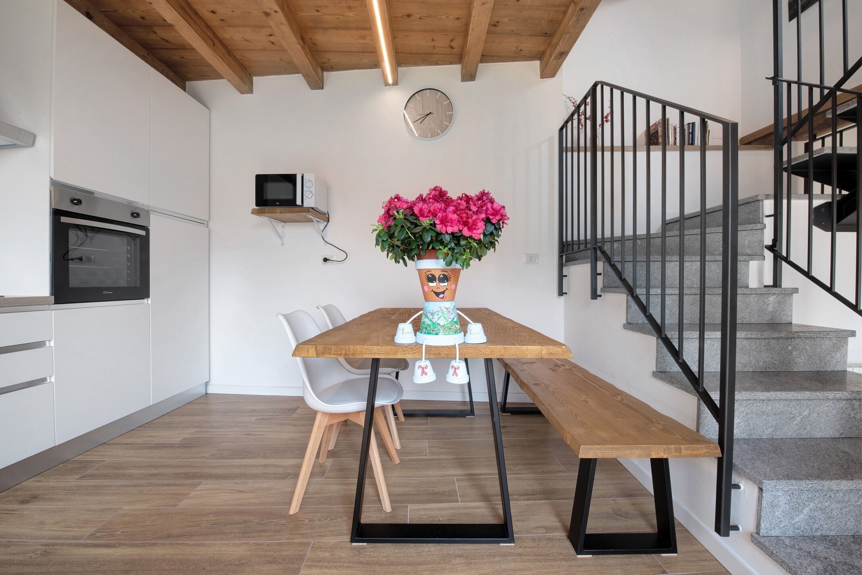 A modern kitchen with a wooden table, chairs, a clock, and a staircase.