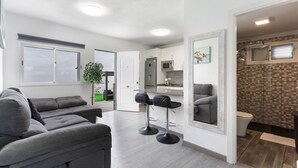 Una sala de estar moderna con un sofá gris, dos taburetes negros, un espejo grande y una cocina con microondas y refrigerador.