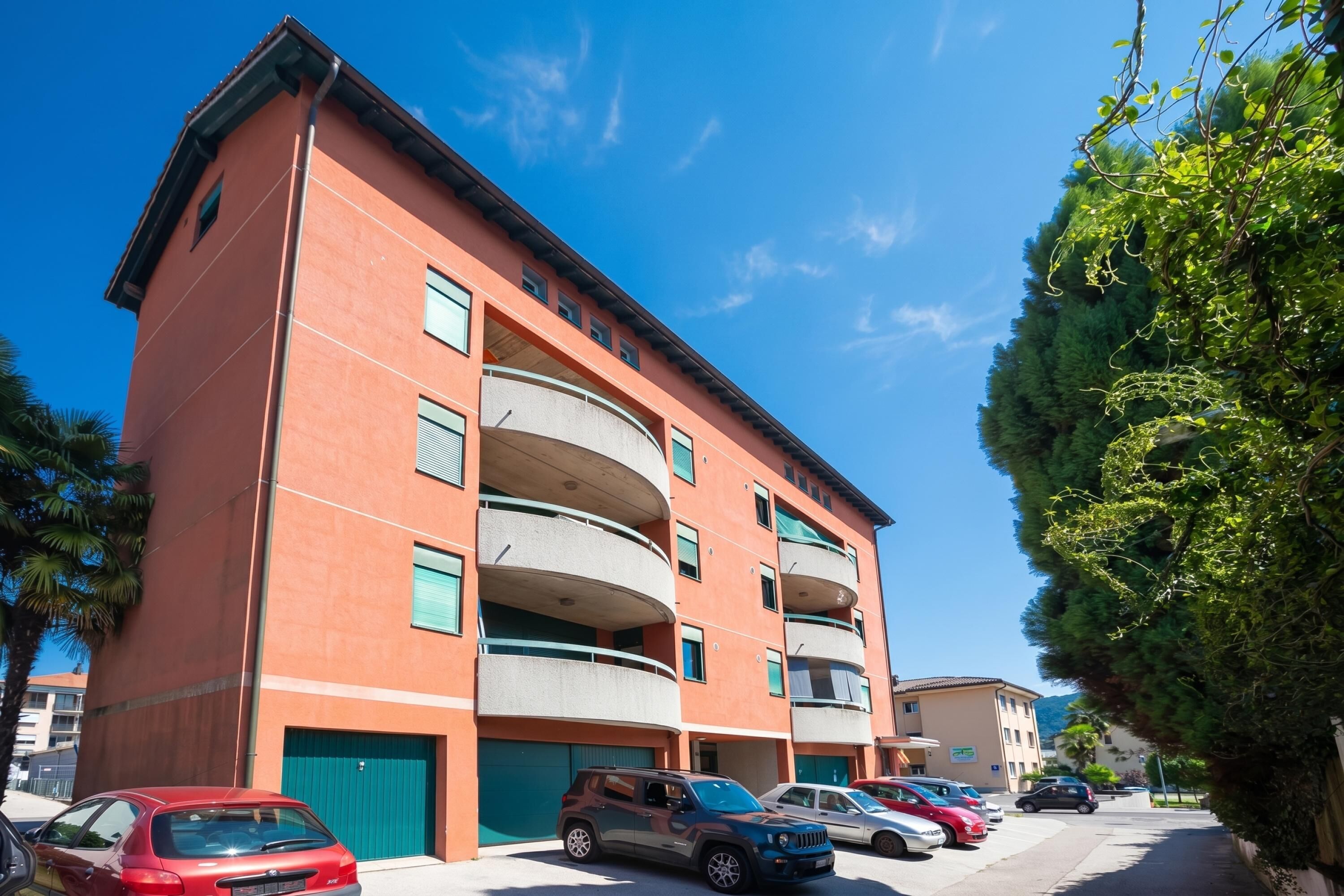 A multi-story building with a parking area in front, featuring a red facade and multiple balconies.