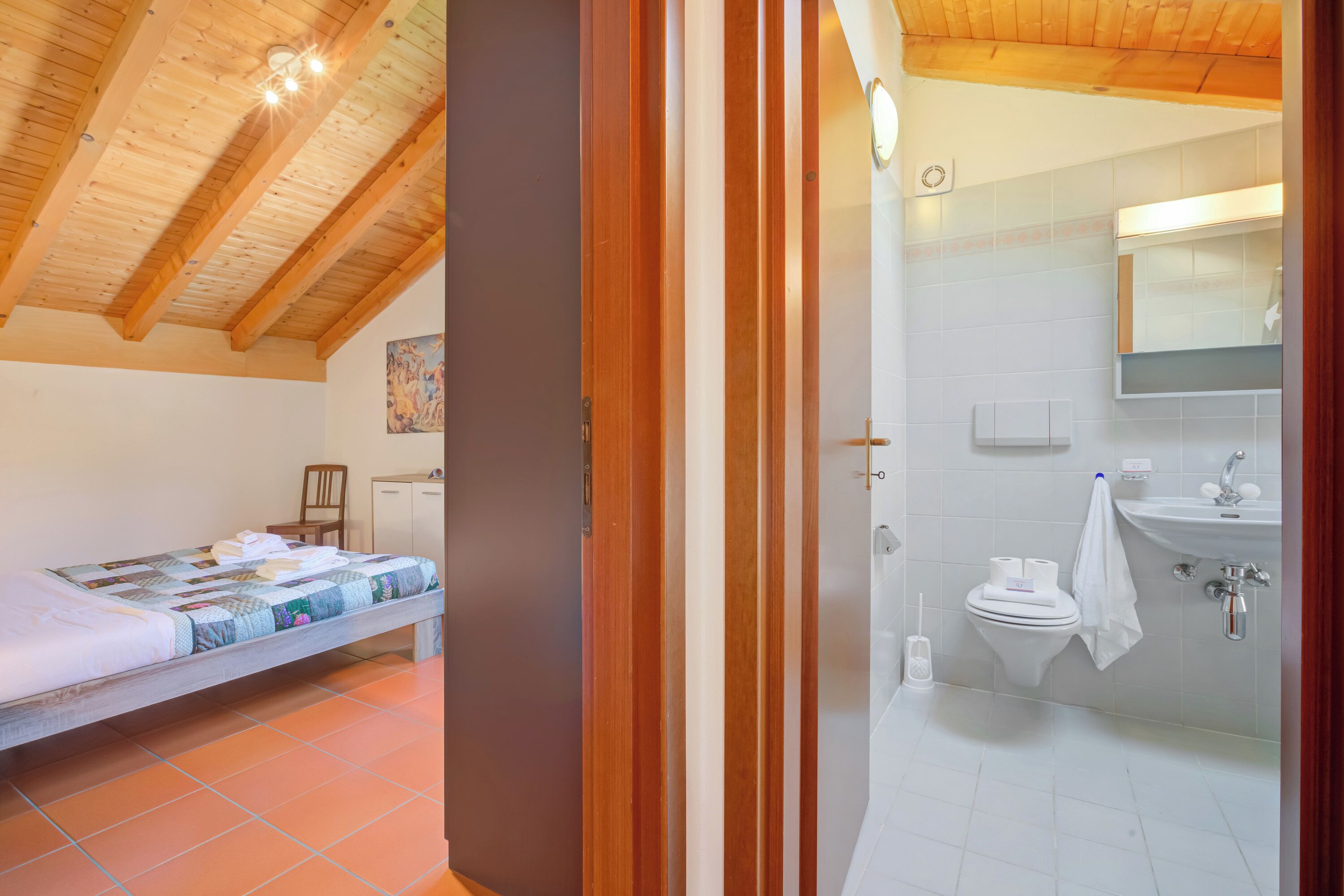 Et soveværelse med træloft, seng med sengetæppe, en stol og et lille skab. Badeværelset har toilet, vask, spejl og et håndklæde hængende på væggen.