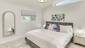 A bedroom with a large bed, a mirror, a window, a ceiling light, and a wall art piece. - Kalispell Gem: Deck, Yard & Fireplace (Kalispell)