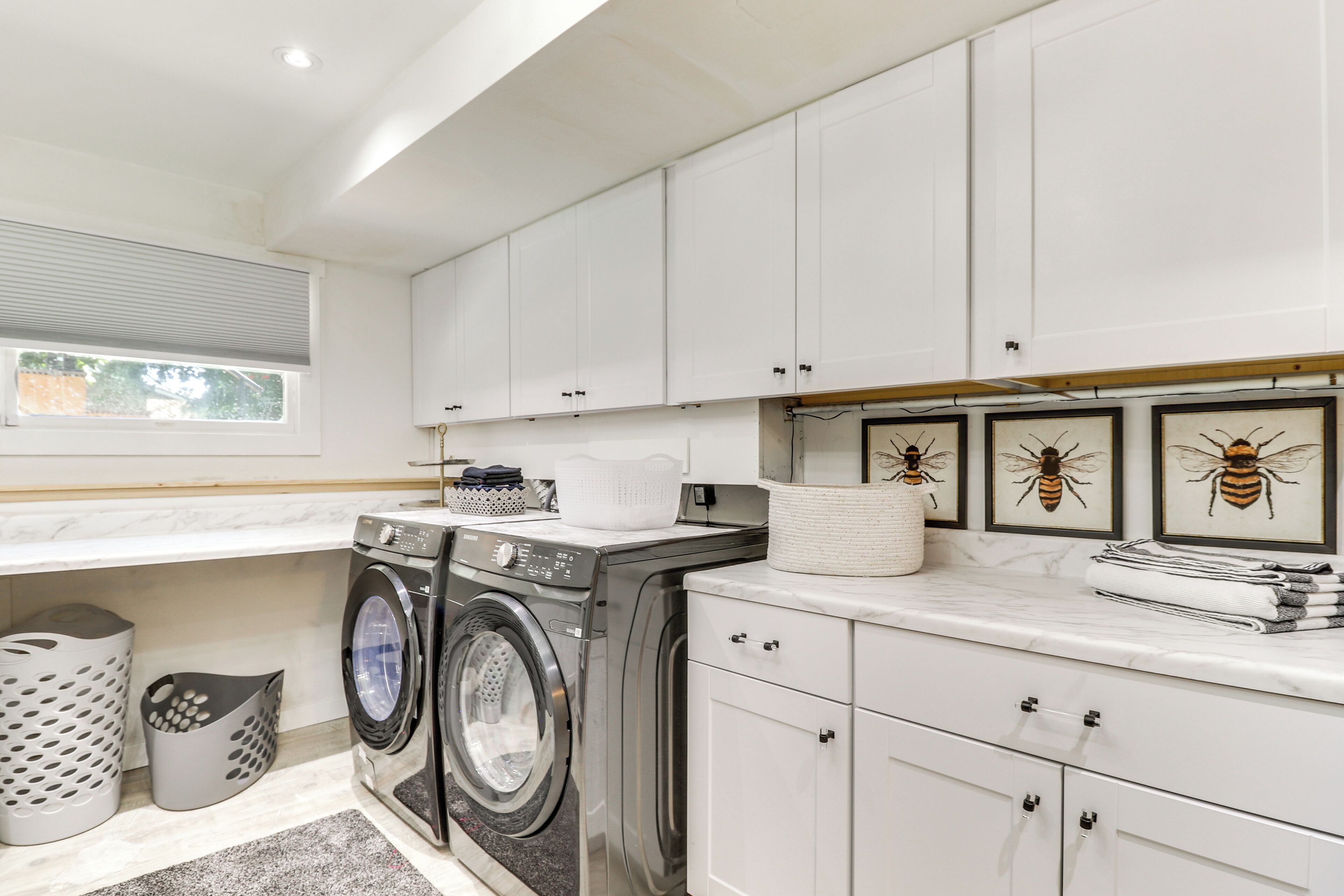 A modern laundry room with two front-loading washing machines, a drying rack, and a sink. There are white cabinets and a window with blinds.