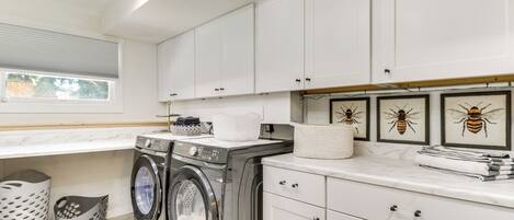 A modern laundry room with two front-loading washing machines, a drying rack, and a sink. There are white cabinets and a window with blinds.