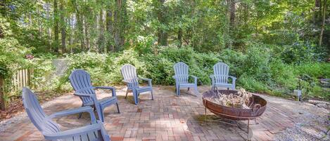 A patio with four Adirondack chairs and a fire pit surrounded by a brick patio, set in a wooded area.