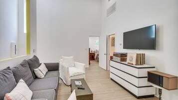 Een moderne woonkamer met een grijze bank, een witte fauteuil en een salontafel. Een flatscreen televisie is aan de muur bevestigd en er staat een houten kast met lades.