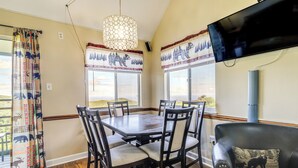 A dining area with a wooden table and chairs, large windows with curtains, a television mounted on the wall, and a leather sofa with a cushion. - 2 Mi to Ober Mountain: Gatlinburg Condo w/ Balcony (Gatlinburg)