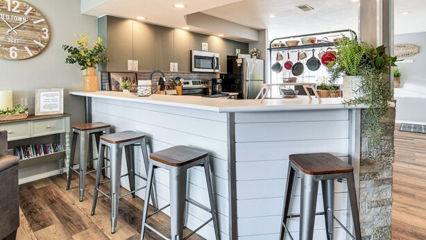 A modern kitchen with a bar area, featuring a large clock, bar stools, and various kitchen appliances. - Condo w/ Pool ~ 2 Mi to Table Rock Lake! (Branson)