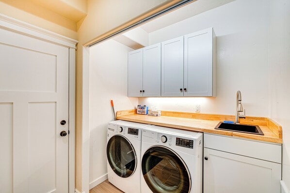 A compact laundry room with two front-loading washing machines, a wooden countertop, a sink, and a cabinet above the machines. - Auburn Farmhouse w/ Fireplace, Close to Wineries (Auburn)
