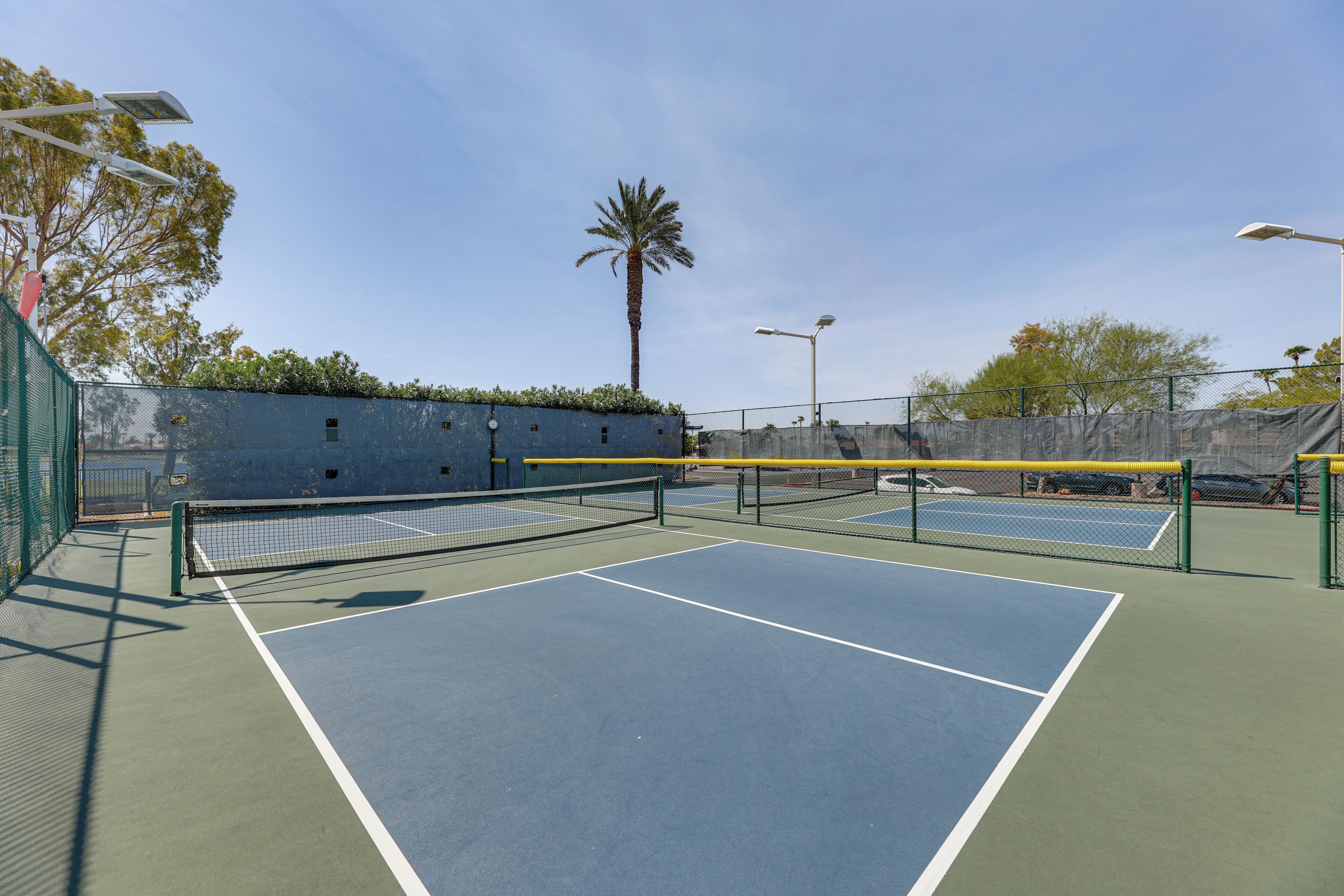 Un court de tennis extérieur avec deux terrains, entouré d’une clôture, et un palmier en arrière-plan.
