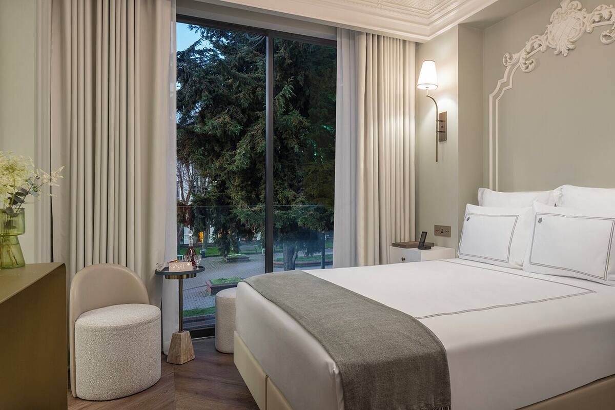 Une chambre d’hôtel avec un grand lit, un bureau, une chaise, une petite table avec un vase et une fenêtre avec des rideaux.