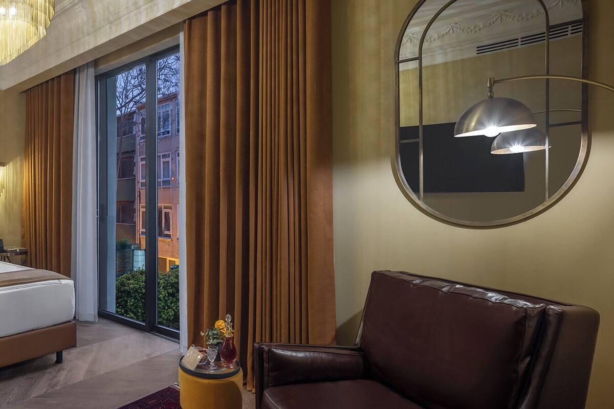 Une chambre d’hôtel avec un grand lit, un fauteuil en cuir, une petite table avec une boisson, et une vue sur les bâtiments par la fenêtre.
