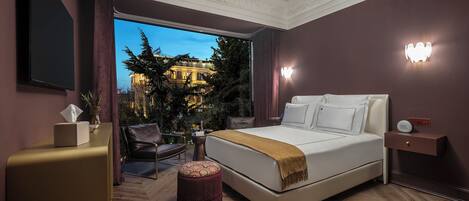 A modern hotel room with a large bed, a desk, a chair, and a view of a building through a large window.