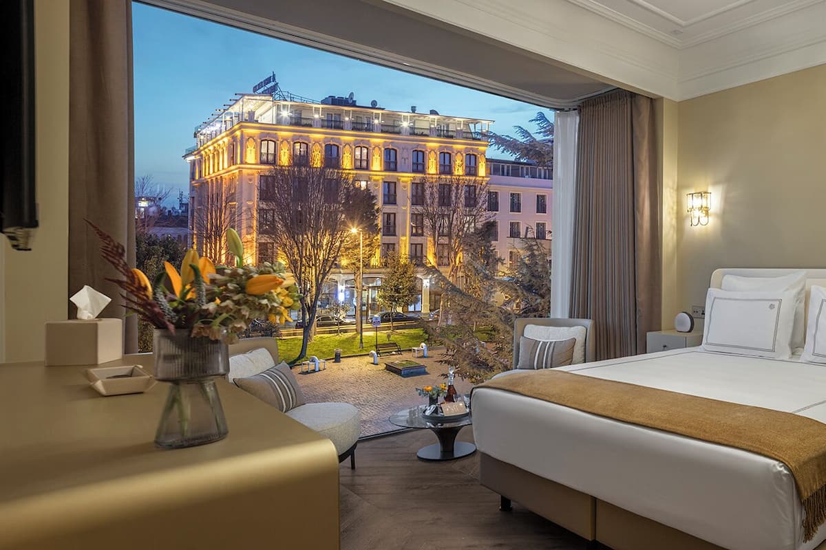 Une chambre d’hôtel avec un grand lit, un coin salon avec une table basse, une vue sur un immeuble situé de l’autre côté de la rue, et un vase rempli de fleurs sur la table de chevet.