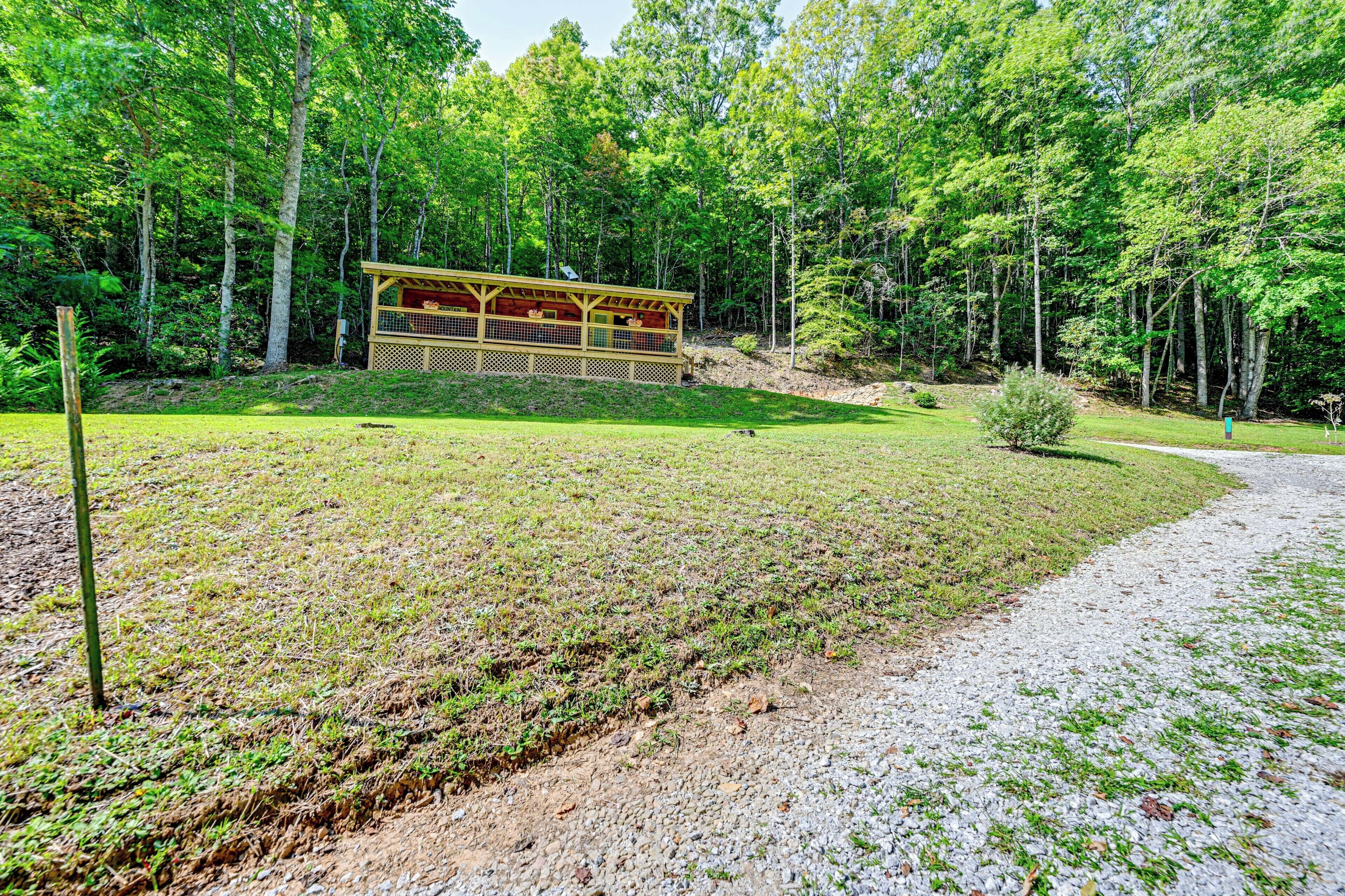 A wooden cabin with a deck, surrounded by a forested area with a gravel path leading to it.