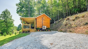 A wooden cabin with a porch, surrounded by a gravel driveway and forested hillside.