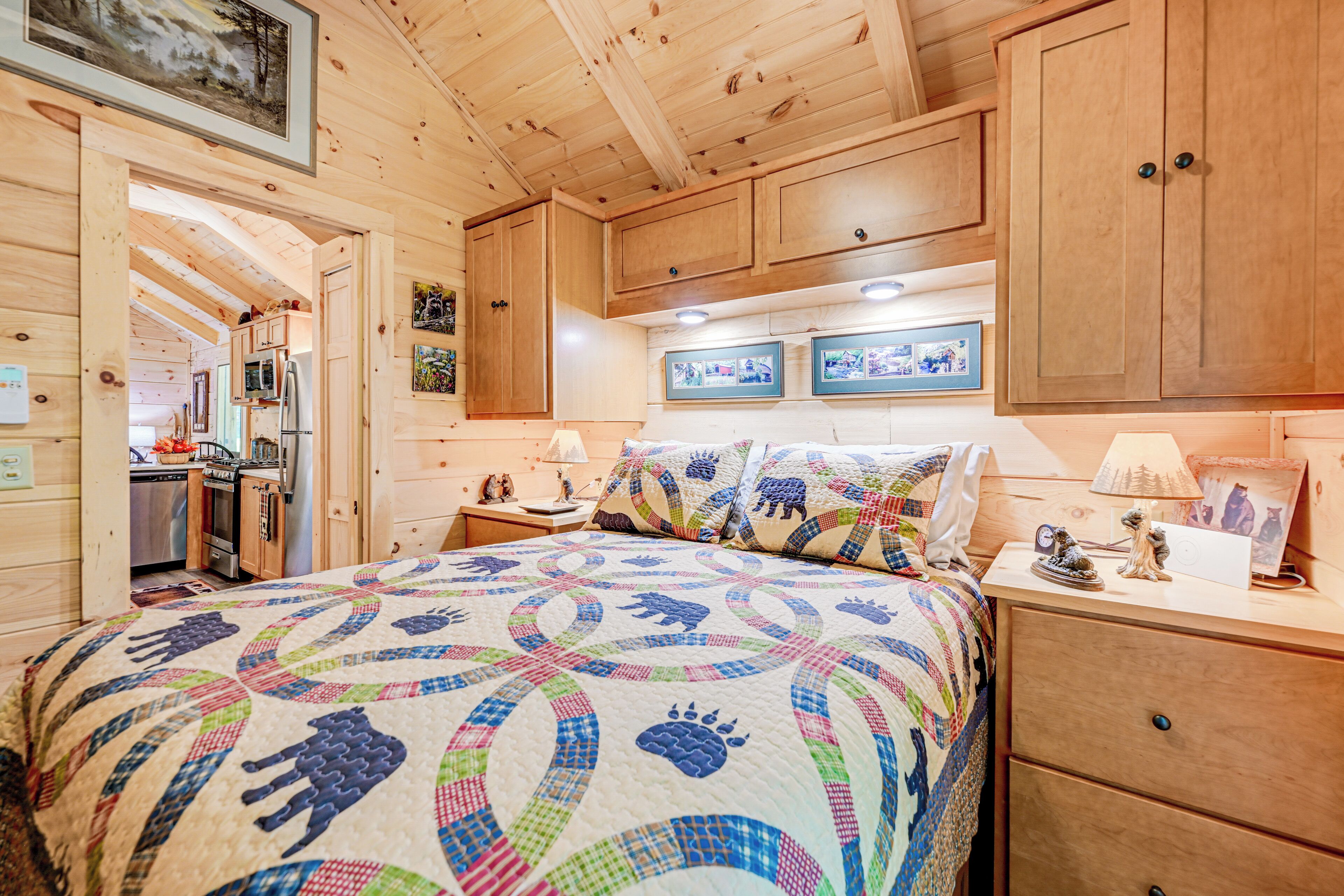 A cozy wooden cabin bedroom with a large bed, wooden cabinets, a small kitchenette, and framed pictures on the wall.