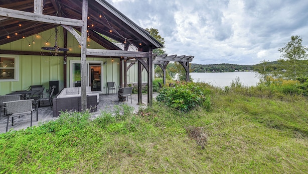 A covered outdoor seating area with wicker furniture and string lights, overlooking a body of water. - Home w/ Private Dock Near Colgate University (Hamilton)