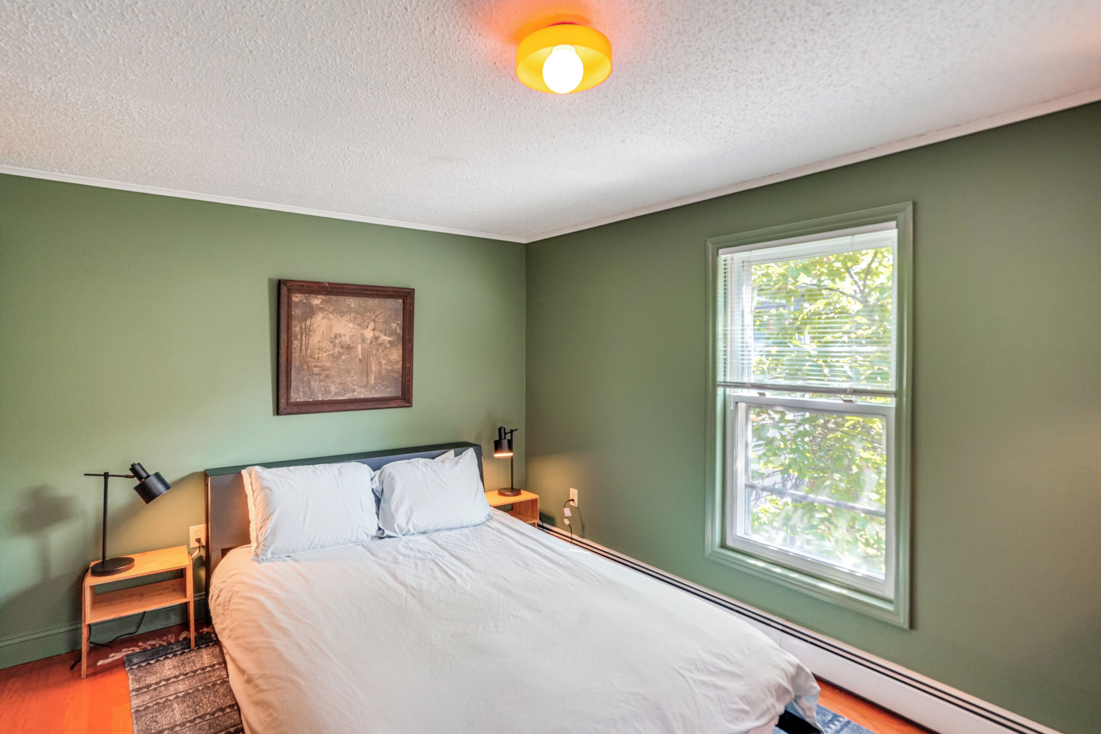A bedroom with a bed, a nightstand, a lamp, a framed picture on the wall, and a window with blinds.