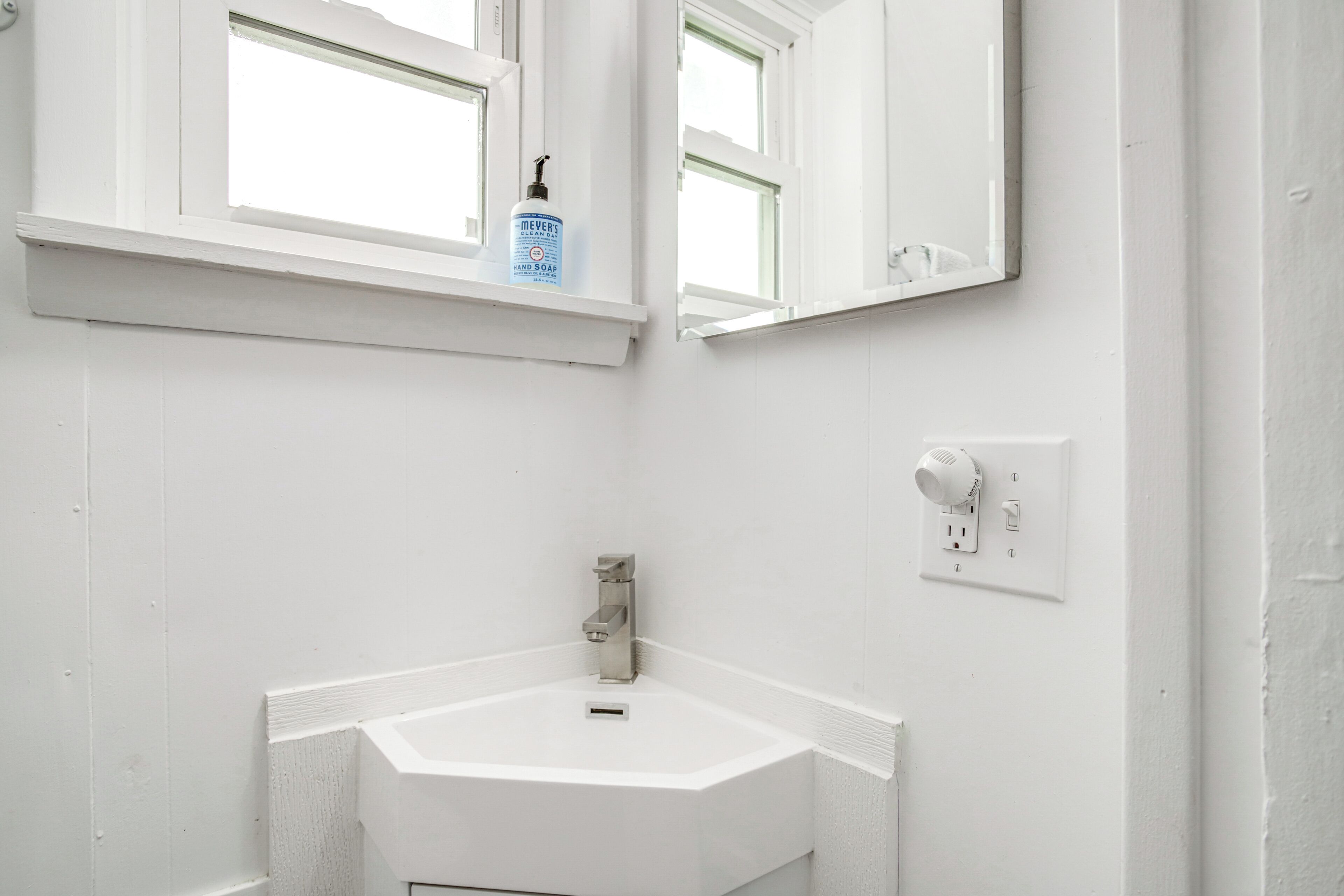 A white bathroom with a corner sink, a window above the sink, and a mirror above the sink.