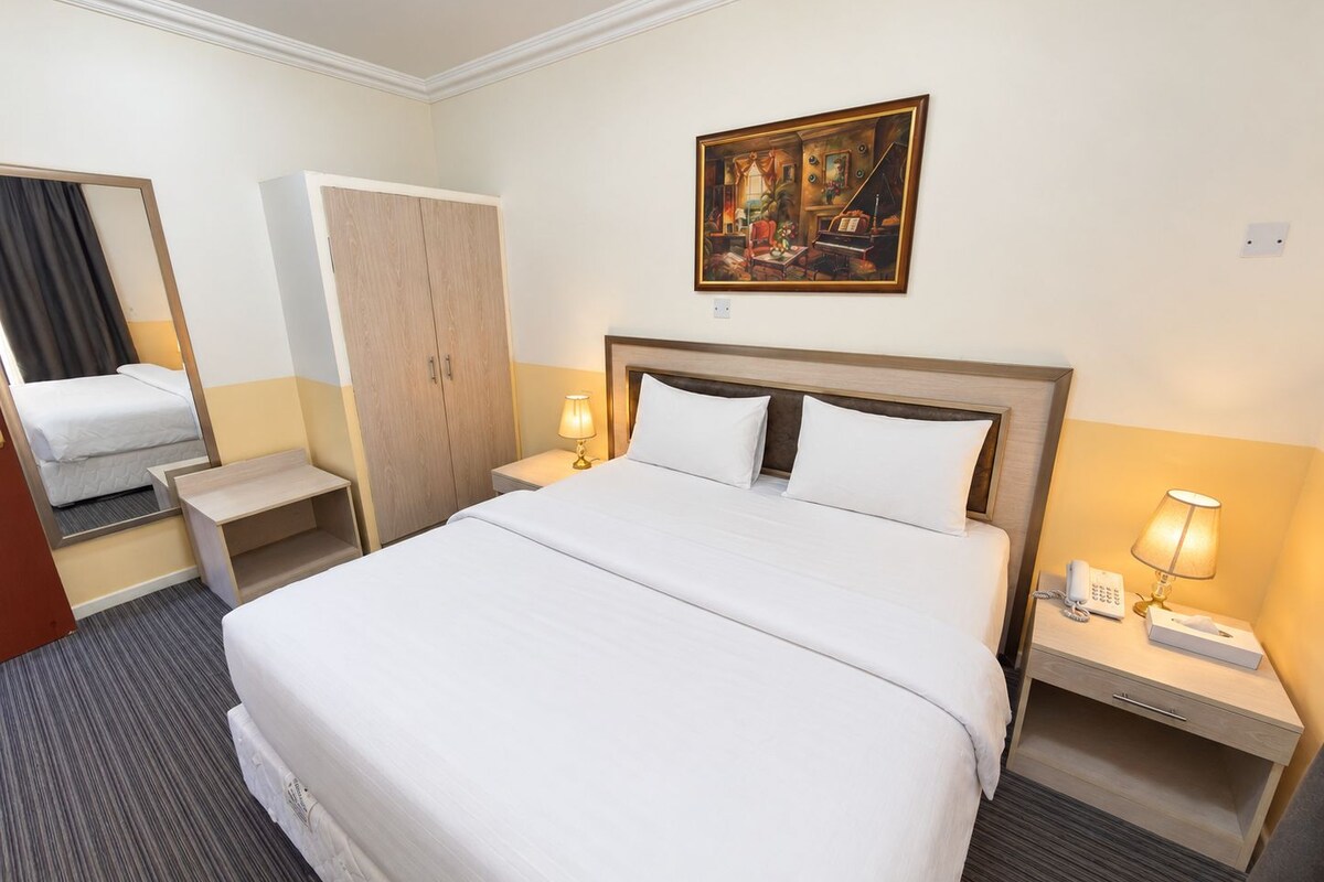 A hotel room with a large bed, bedside tables with lamps, a wardrobe, a painting on the wall, and a mirror.