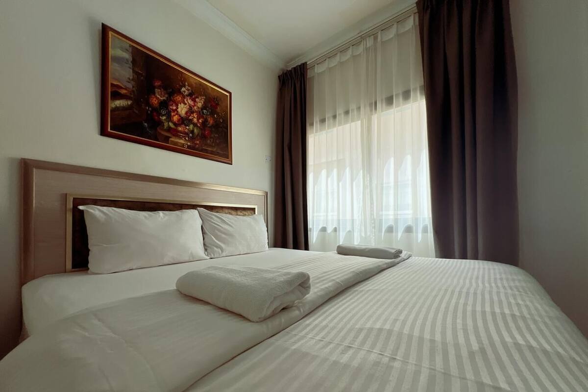 A neatly made bed with white linens and a striped blanket, a wooden headboard, a painting of flowers on the wall, and a window with sheer curtains.