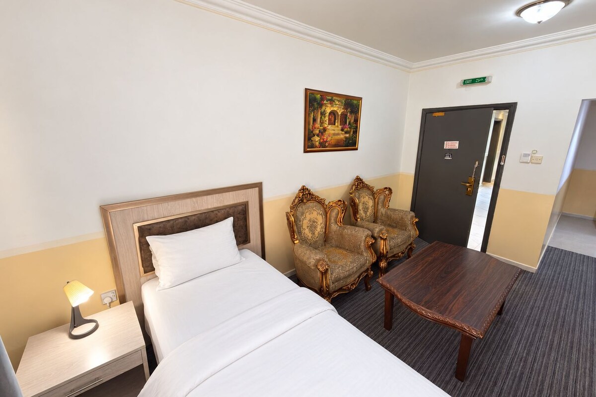 A hotel room with a bed, two ornate armchairs, a small wooden table, a nightstand with a lamp, and a painting on the wall.