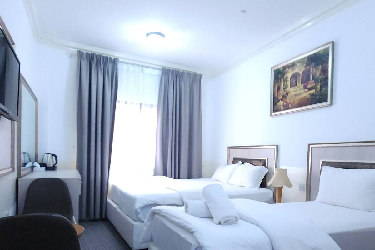 A hotel room with two beds, a desk, a chair, a painting on the wall, and a window with curtains.