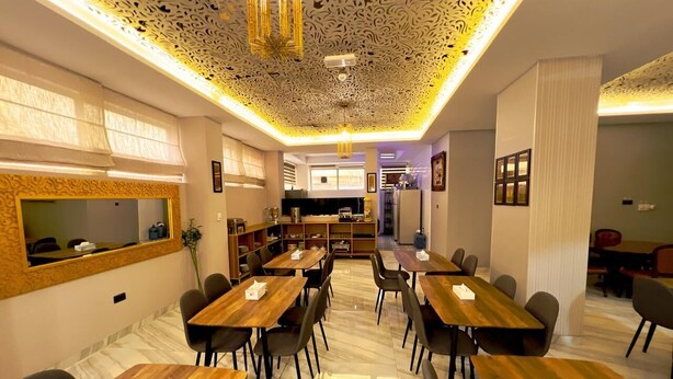 A modern dining area with wooden tables and chairs, a decorative ceiling, and a kitchen in the background.