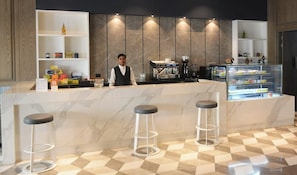 A modern hotel lobby with a marble reception desk, bar stools, a coffee machine, and a display case with pastries. - Evanthe Hotel (Lucknow)