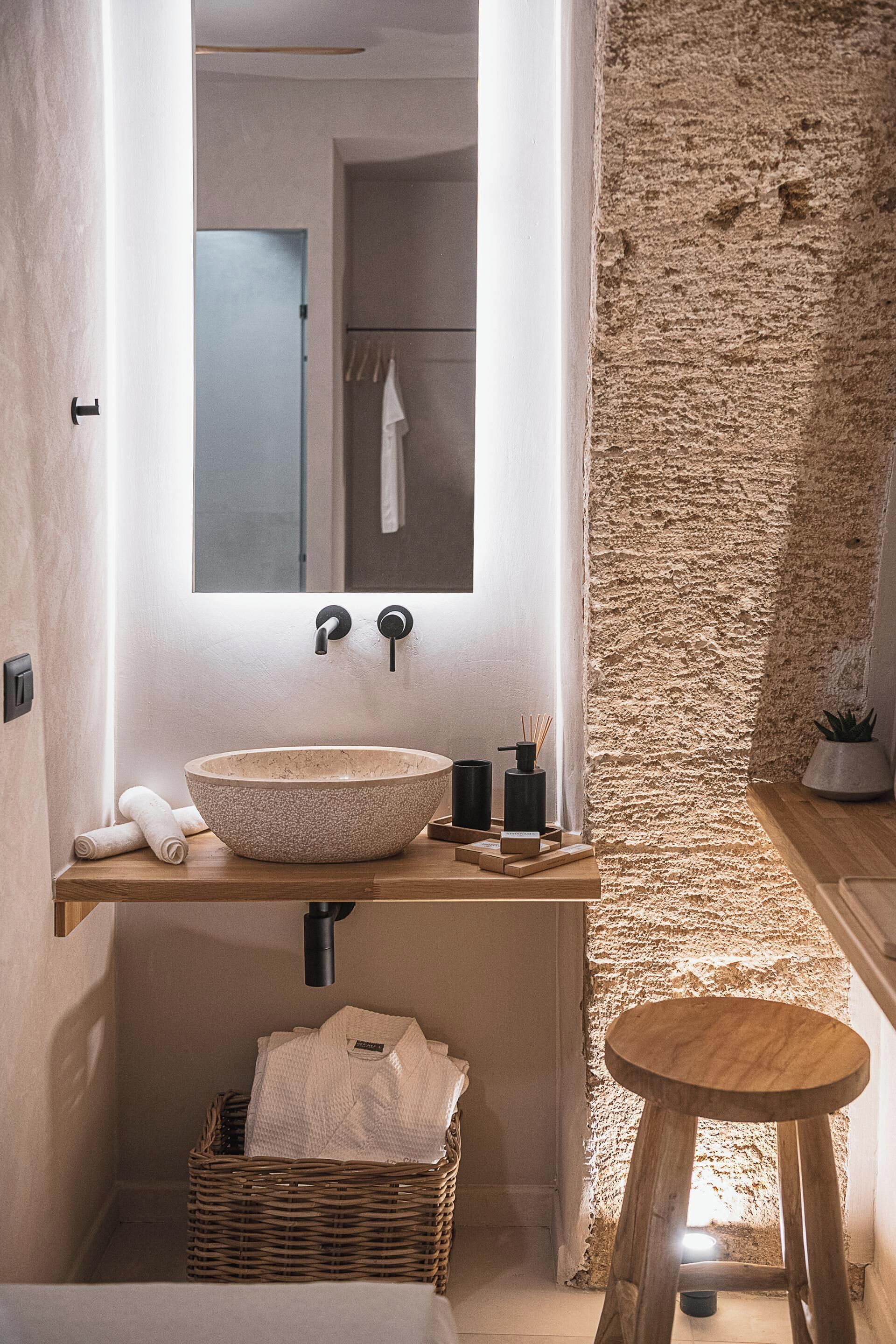 Een badkamer met een grote stenen muur, een houten wastafelmeubel, een rond krukje en een rieten mand.