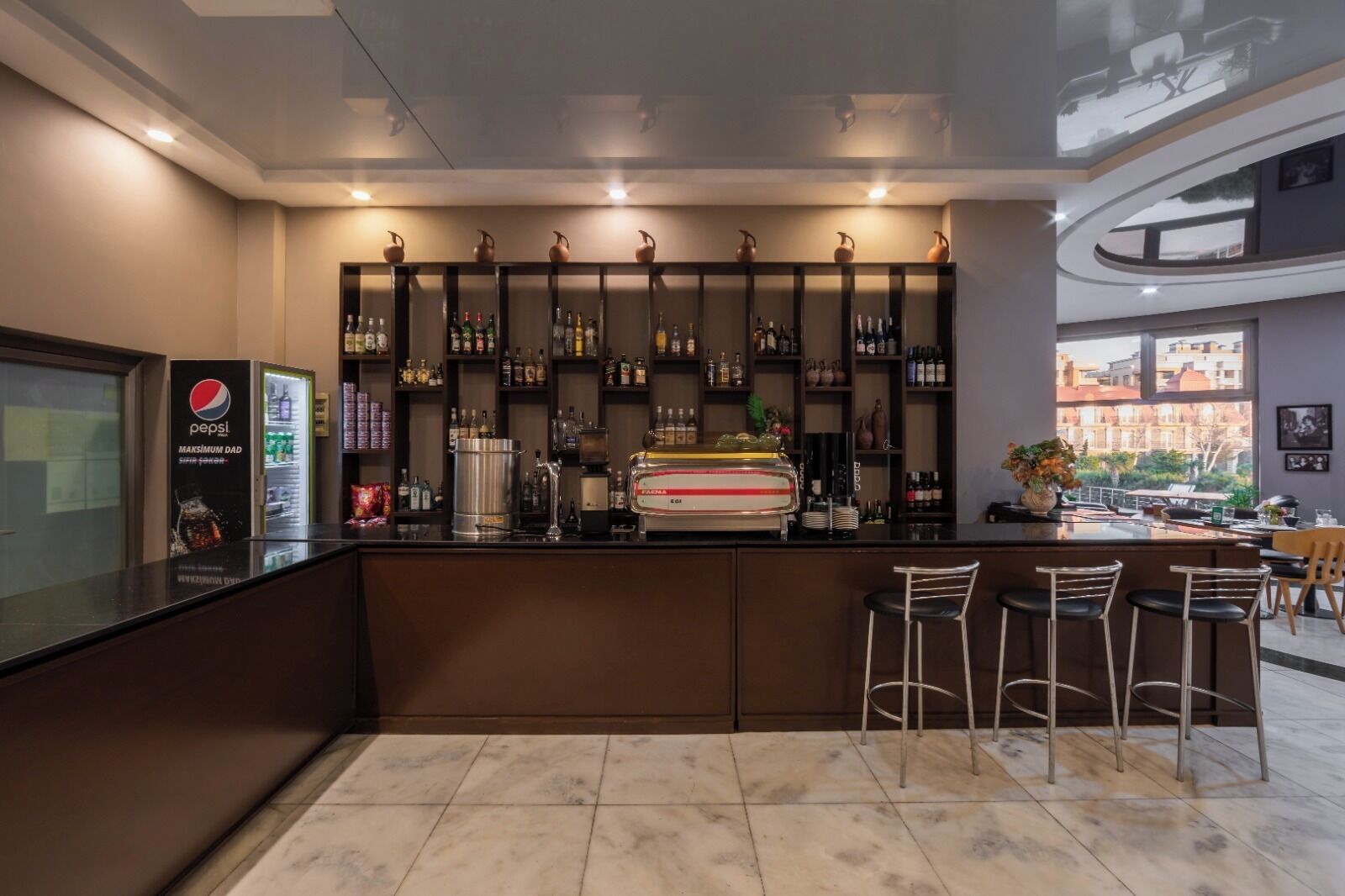 Uma área de bar com balcão, prateleiras de garrafas, refrigerador de Pepsi e vista para prédios lá fora.