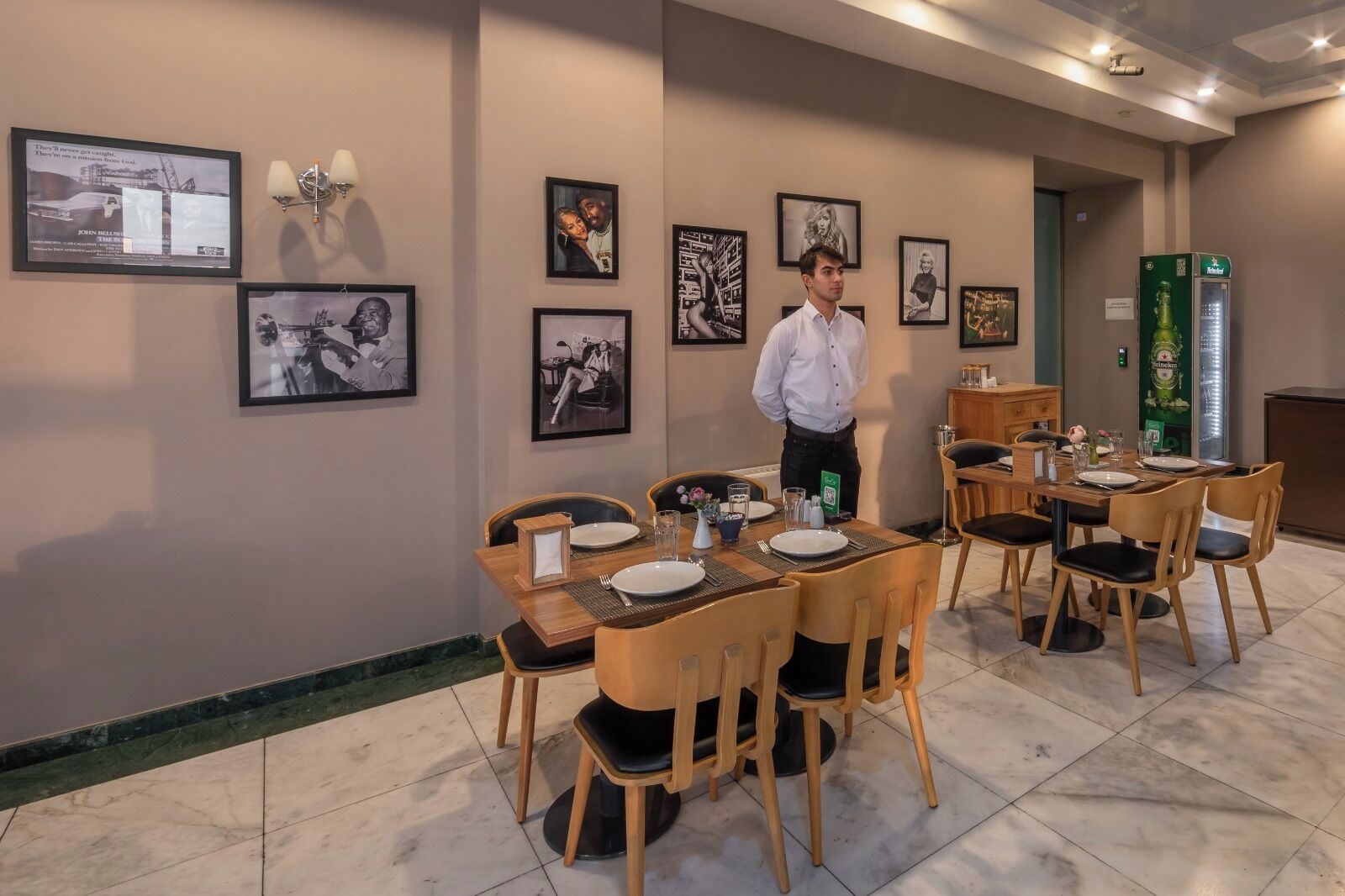 A dining area with tables set for four, framed pictures on the walls, and a refrigerator with a Heineken logo.