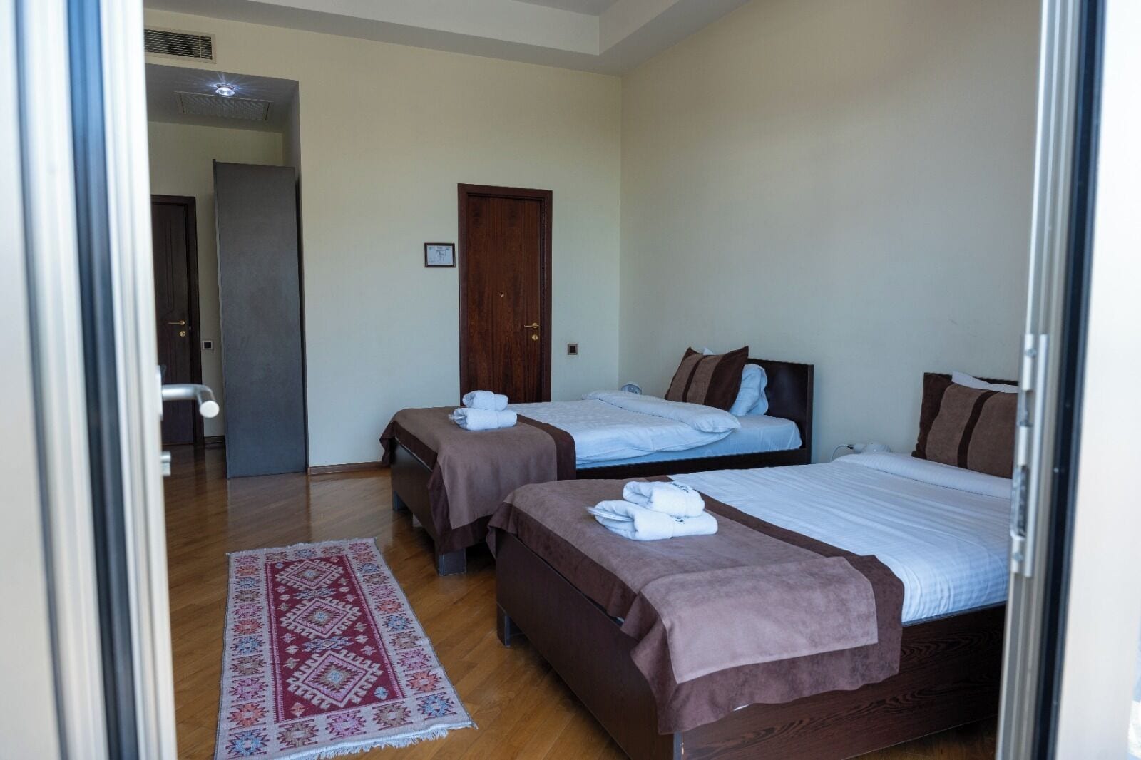 A hotel room with two beds, a wooden floor, a red patterned rug, and a door leading to another room.
